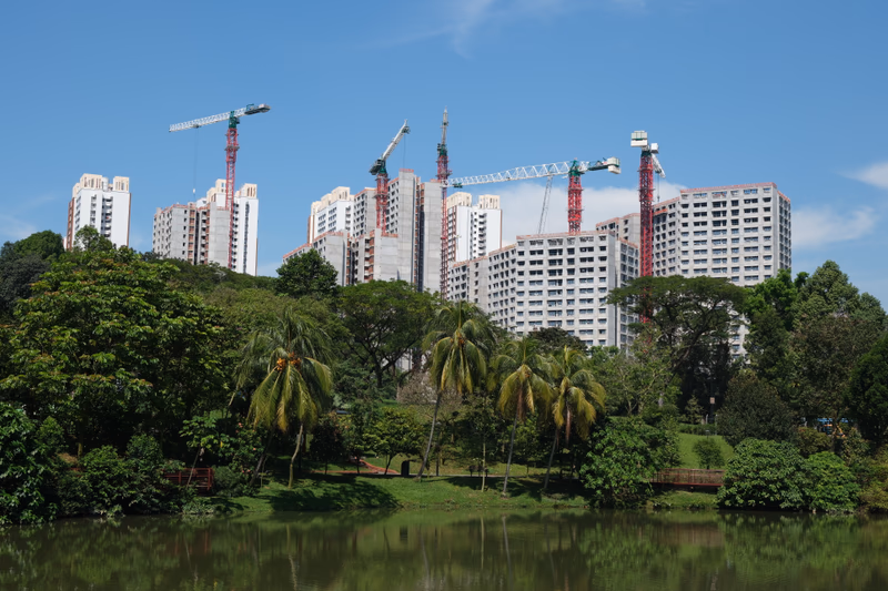 Aerial view of a residential area with multiple high-rise buildings under construction, surrounded by lush greenery and a serene body of water.